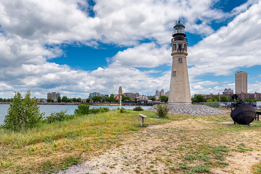 About Our Agency - Buffalo Lighthouse Park in Buffalo, NY, Water and Skyline in the Background, a Grass Sandy Area in the Foreground, White Puffy Clouds Overhead in a Deep Blue Sky