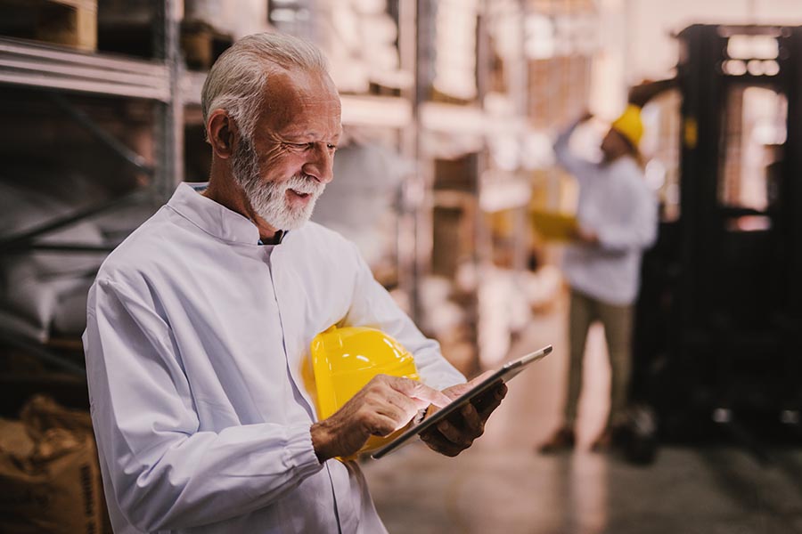 Client Center - Senior Businessman Uses a Tablet in a Warehouse, Yellow Hard Hat Under His Arm